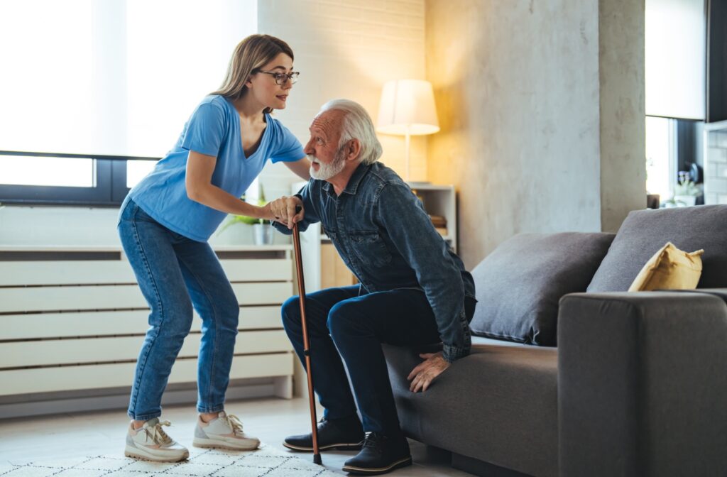 A woman caregiver helping an older adult man get up off a couch from a seated position.