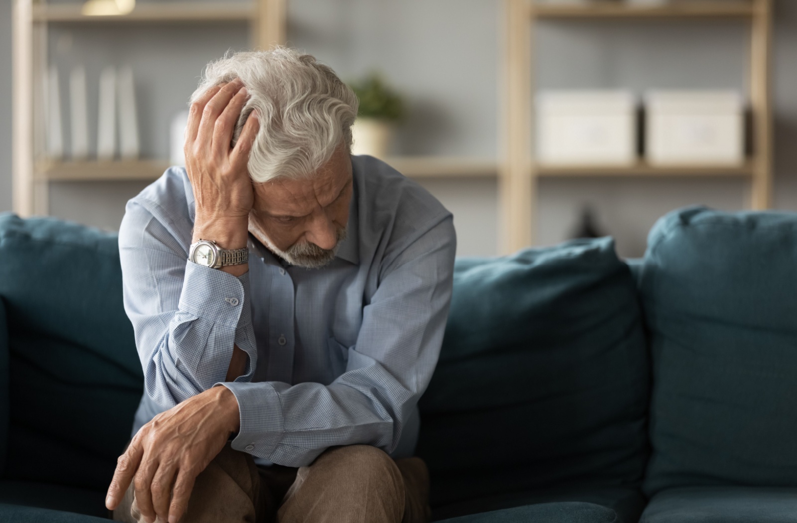 An older adult man with dementia sits on a blue couch with one hand on his head looking worried.