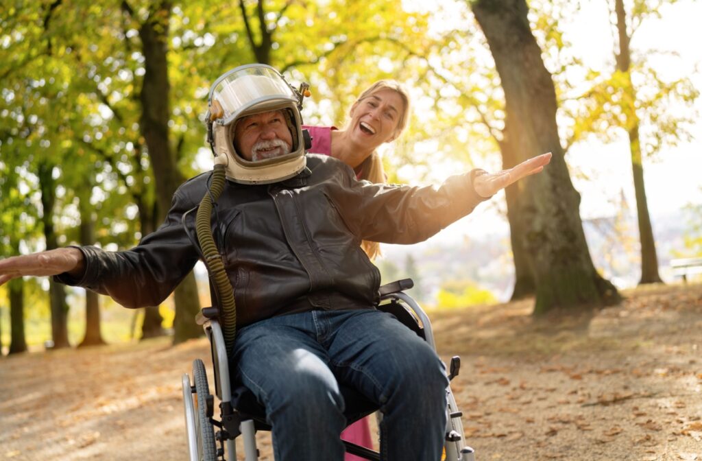 An older adult man in a wheelchair wears an astronaut helmet as a smiling nurse in pink scrubs pushes him through a park.