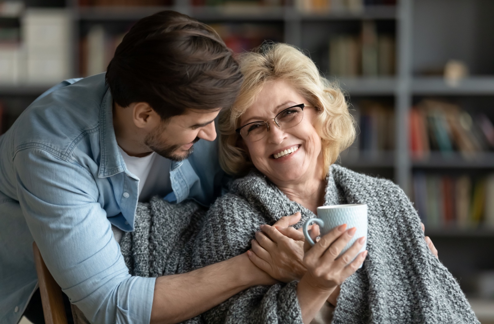 Older woman with a blanket around her shoulders and holding a mug of coffee smiles at her adult son who hugs her from behind.