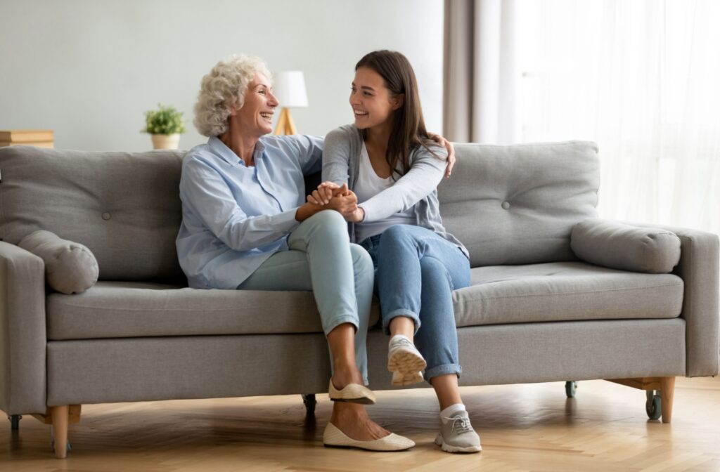 An adult daughter sitting on a couch with her senior mother while they hold hands and smile at each other.