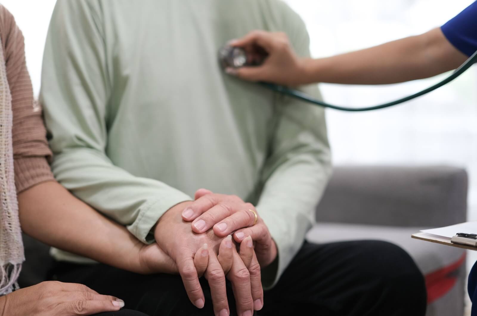 A close-up an older adult holding hands with an adult child while a healthcare professional checks his chest with a stethoscope.