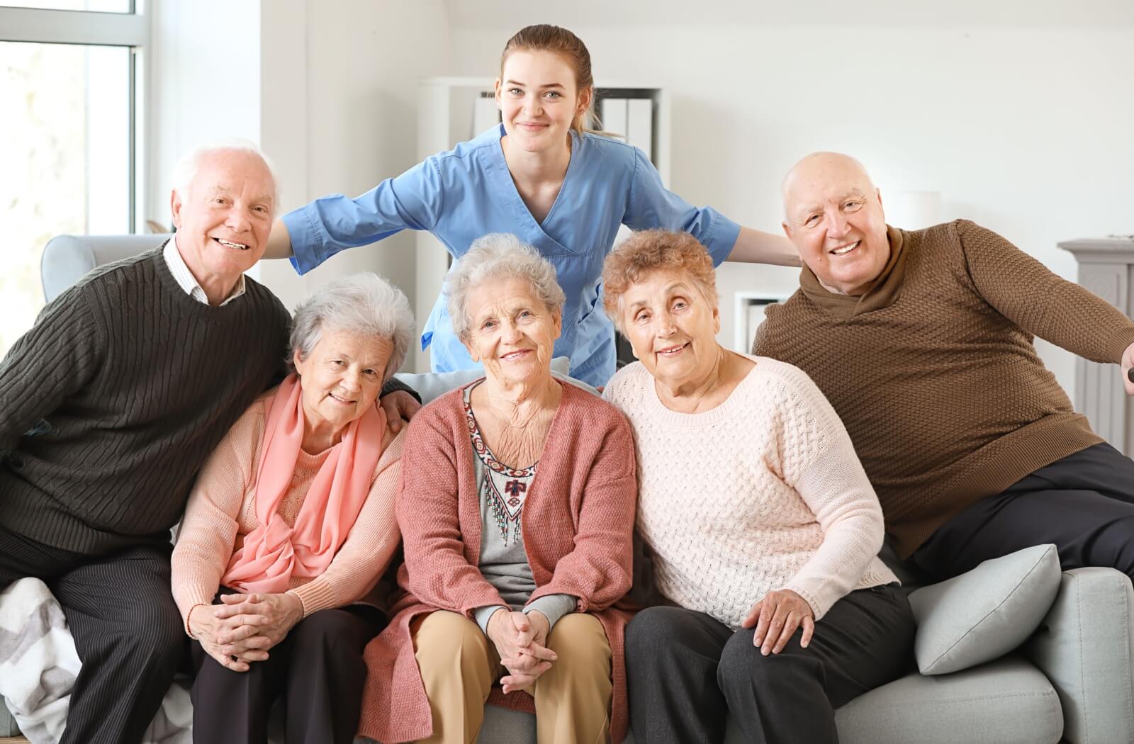 A group of older adults in senior living sitting together on a couch in front of a smiling caregiver.