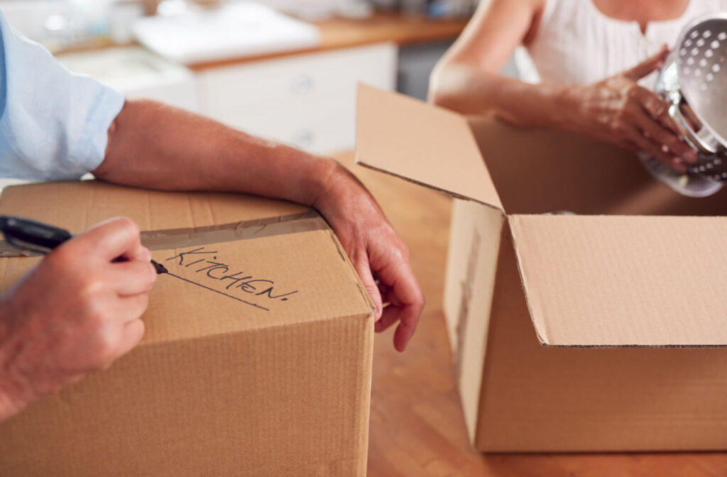 A close-up image of an older adult writing the word "Kitchen" on a box while packing to move to assisted living.