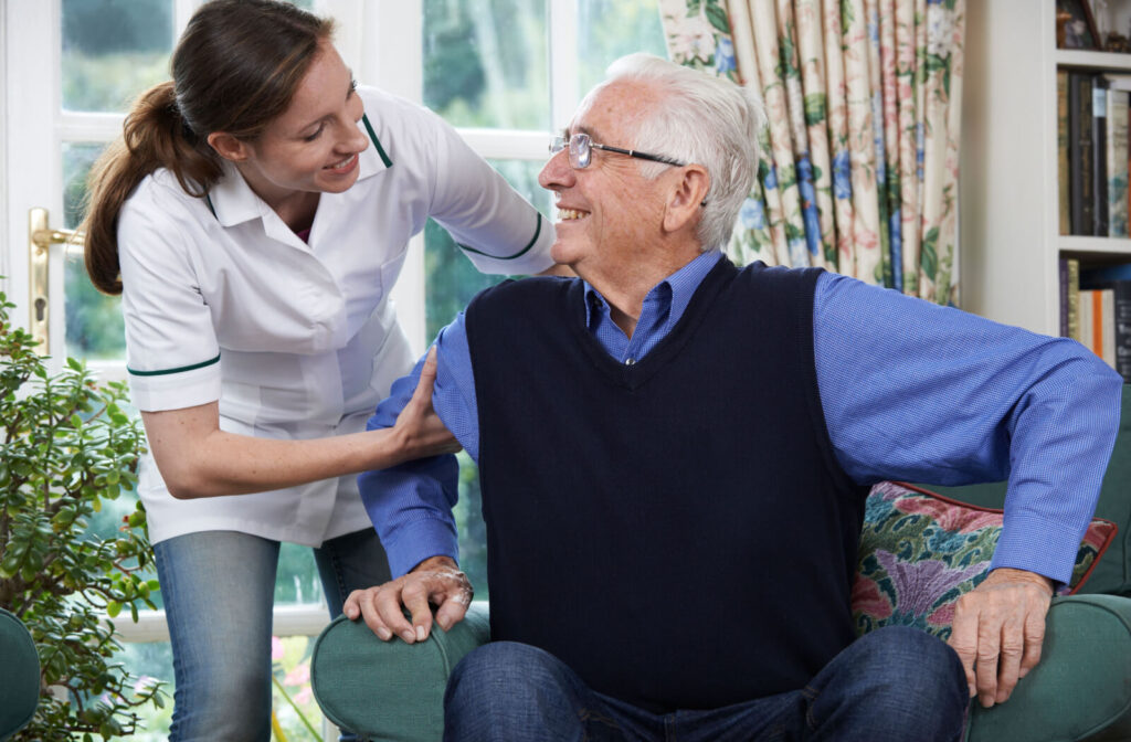 A caregiver helps an older adult stand while they smile in appreciation in a library in an assisted living community.