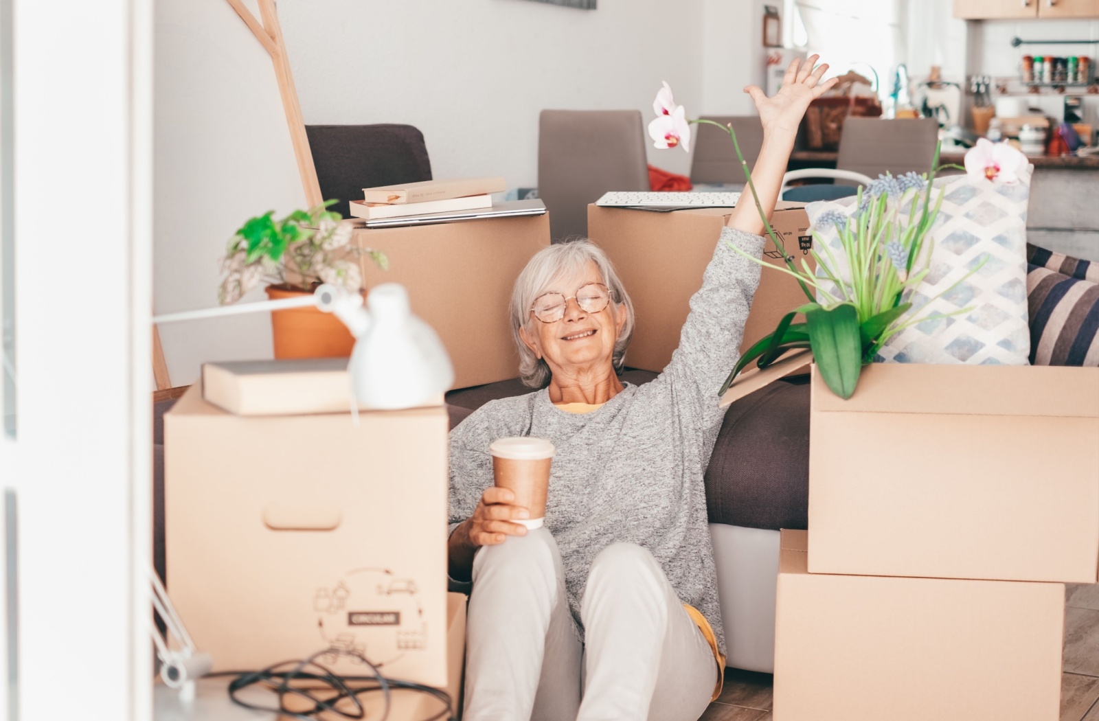 An older adult sitting on the ground with a coffee, surrounded by packed-up boxes for a move to assisted living.