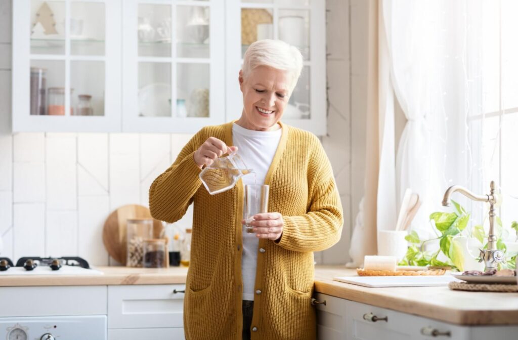 A happy senior in a yellow cardigan pours themselves a glass of water to stay hydrated throughout the day