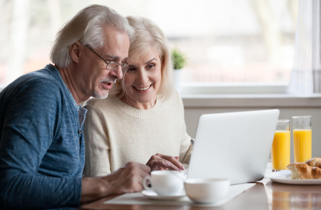An older adult shows their spouse a potential senior living community on their laptop over breakfast at their kitchen table
