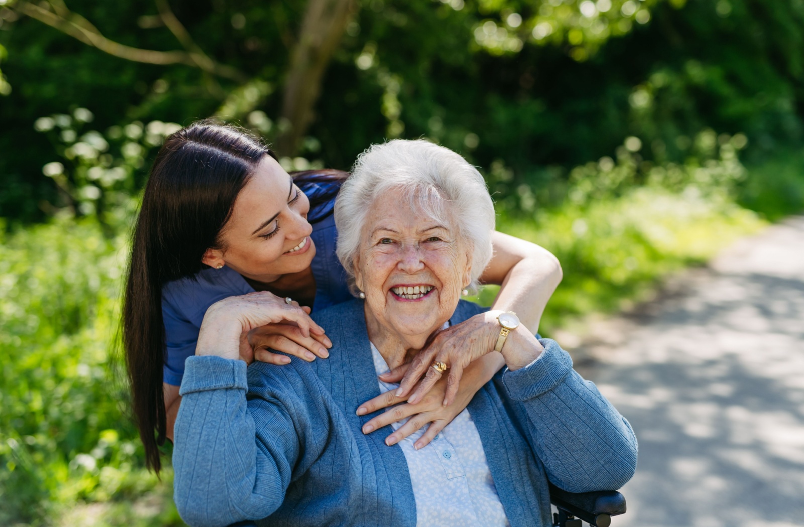 A caregiver embraces a senior from behind outdoors on a sunny day surrounded by greenery